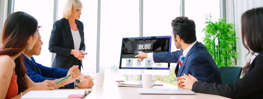 Business team in a meeting with a man pointing at a computer screen displaying a blog page
